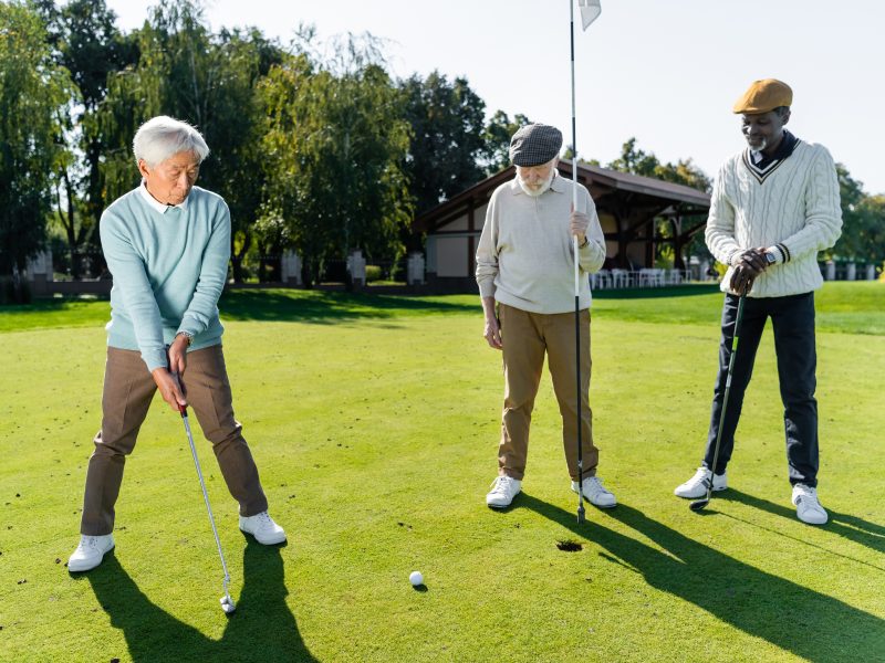 senior man in flat cap holding flag stick near hole and multiethnic friends