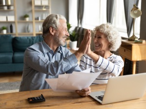 Overjoyed exited middle aged married couple giving high five, finishing doing domestic paperwork together at home. Euphoric happy older mature spouses celebrating successful investment or purchase.